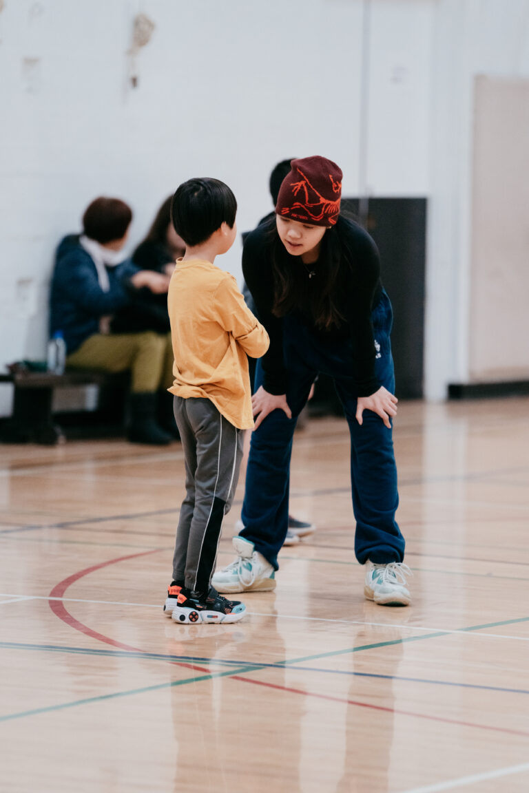 A young player in a yellow shirt stands on a gym court as a coach in a red beanie bends down to talk with him