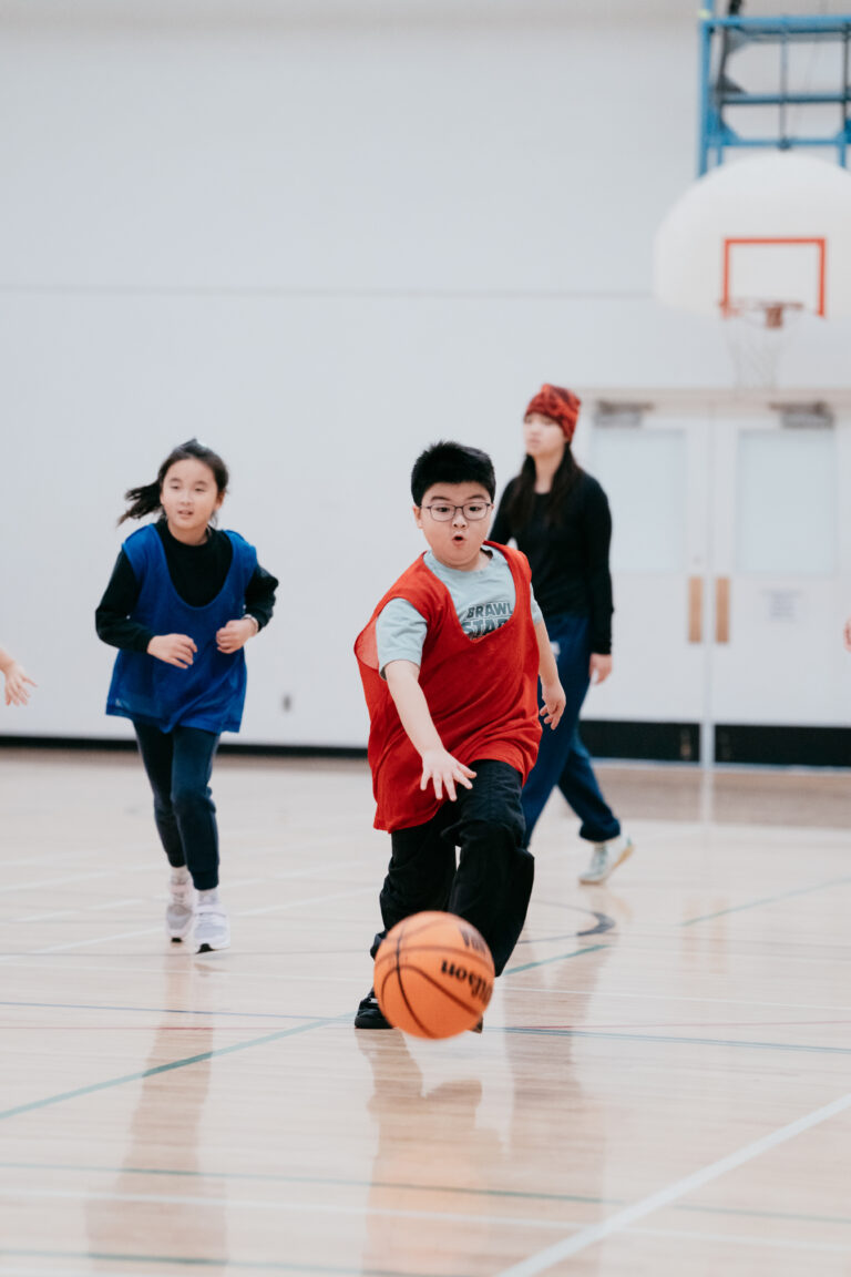 A boy in a red pinnie dribbles a basketball up a gym court as other players and a coach follow