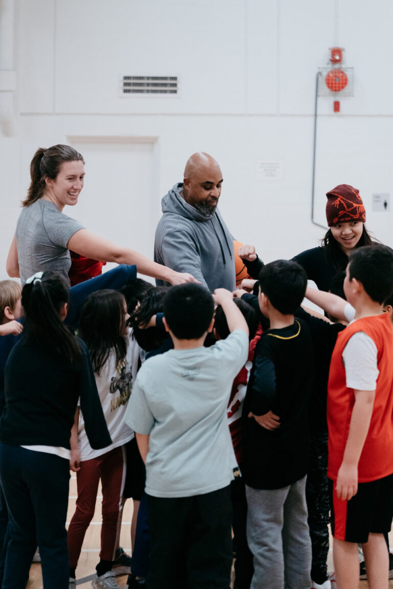 Coaches and young players join hands in the middle of a team huddle in a gym