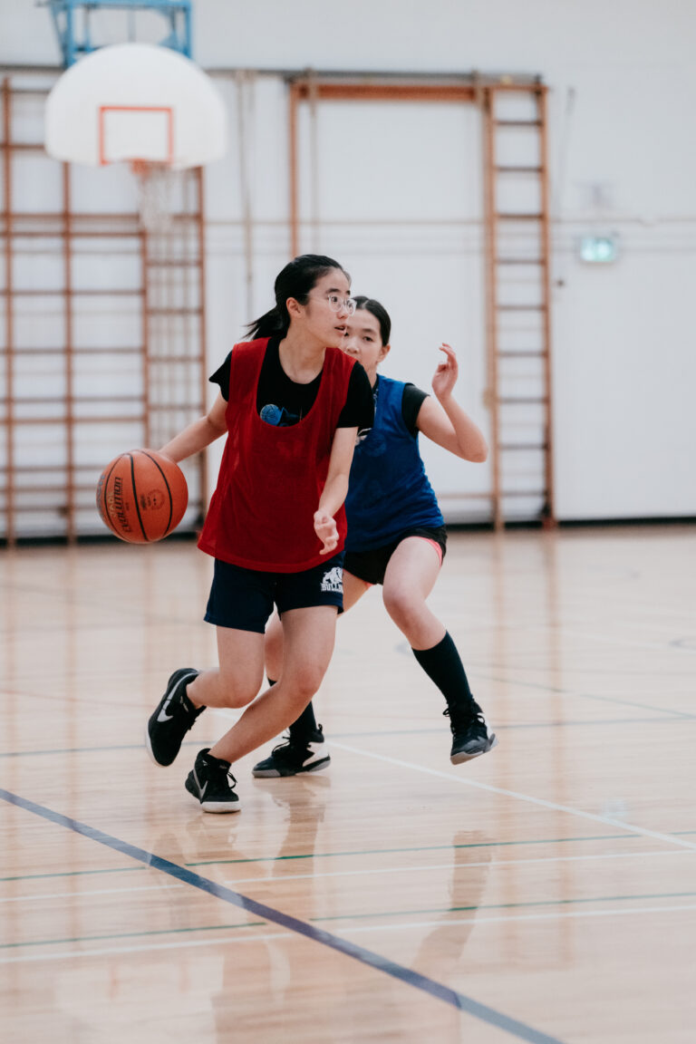 A girl in a red pinnie dribbles a basketball while a player in blue defends her in a gym